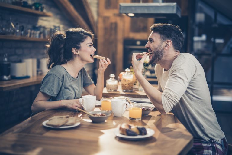 Young happy couple having fun while eating sandwiches for breakfast in the kitchen.